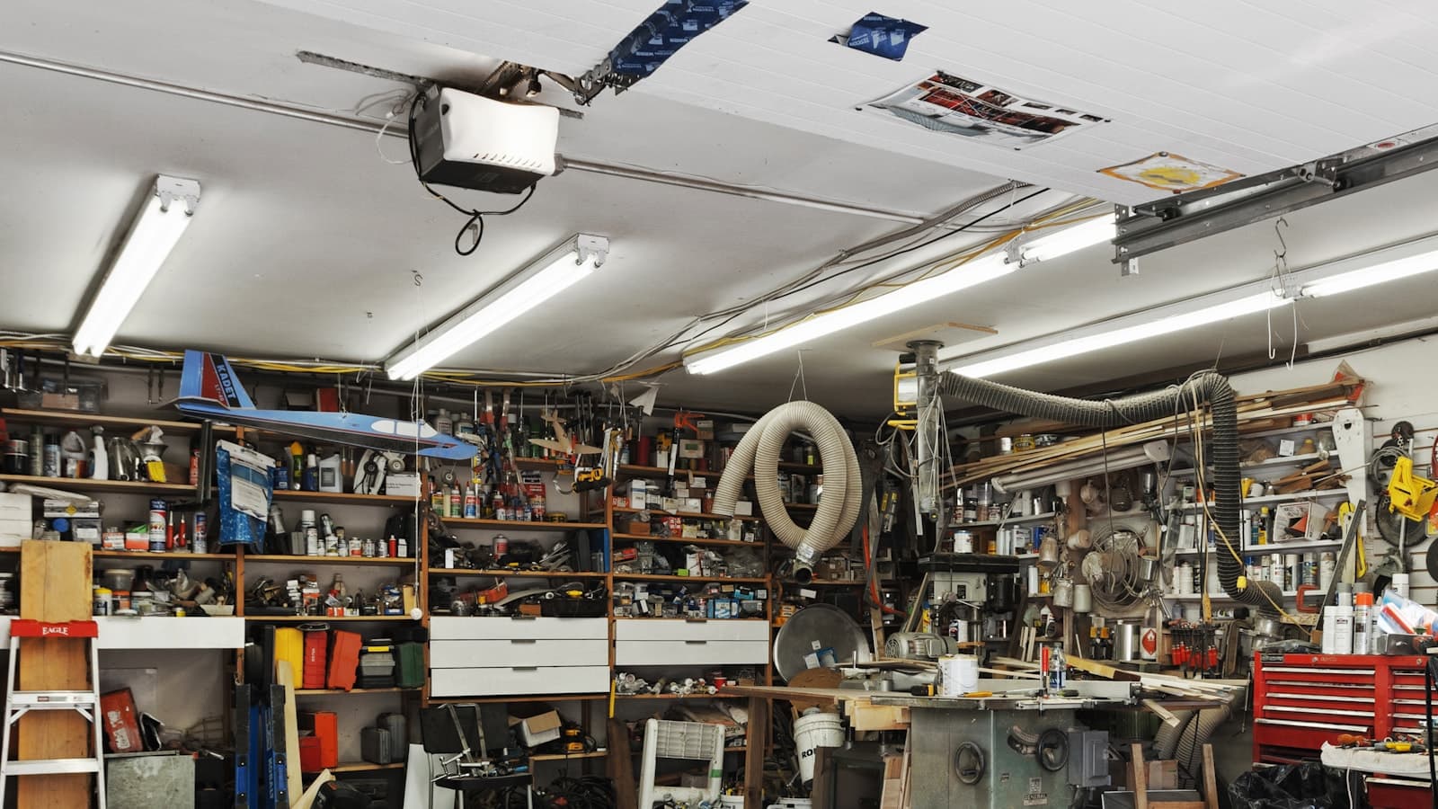 Cluttered garage with shelves of tools, boxes and stored items lining the walls above a concrete floor