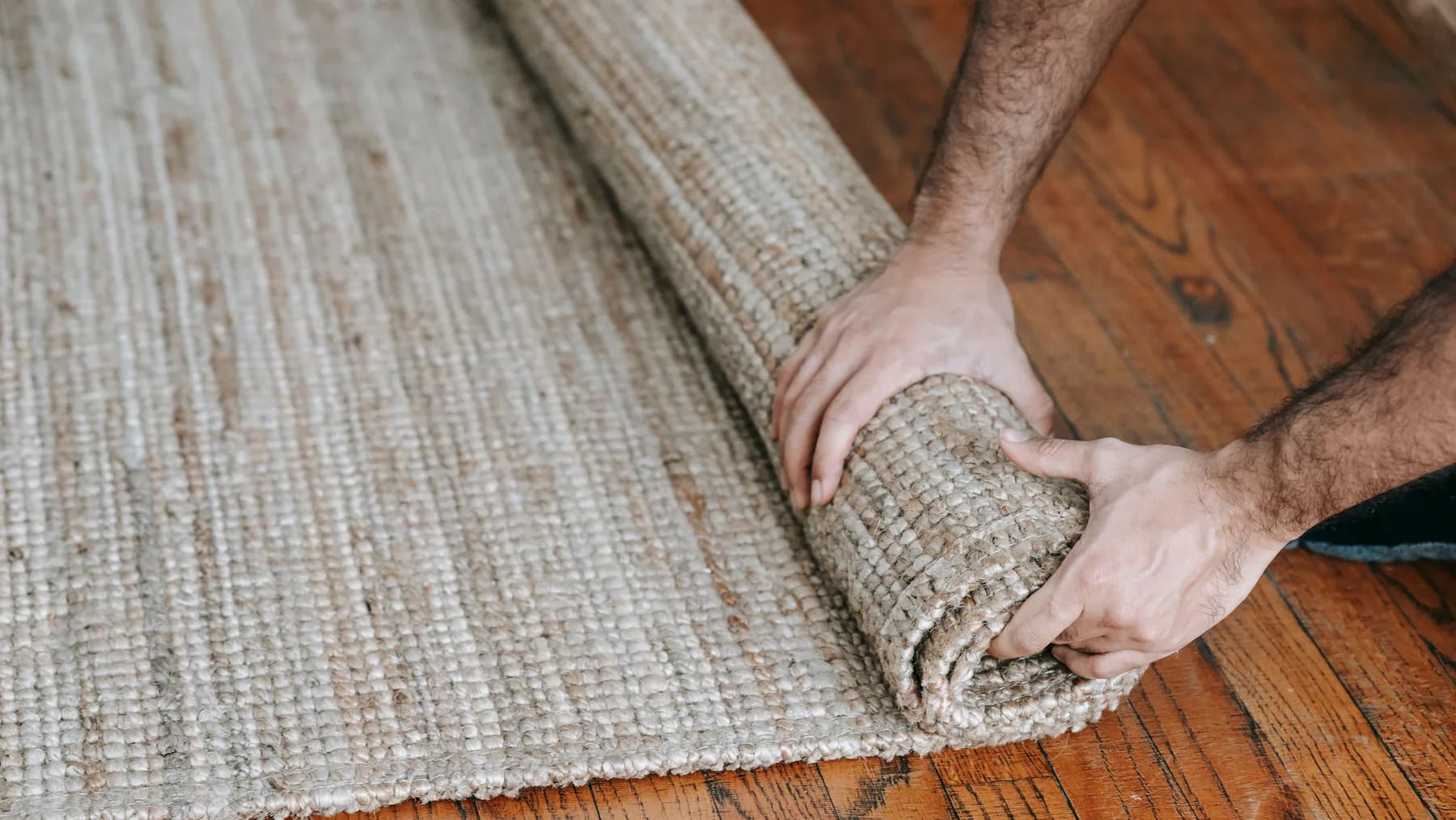 Two hands rolling up a woven jute floor rug from one edge on a wooden floor