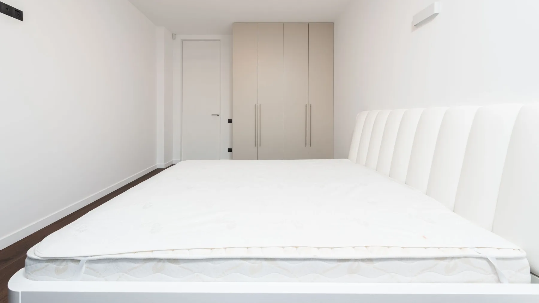 Bare white mattress with a quilted topper on a low bed frame in a plain bedroom with a fitted wardrobe