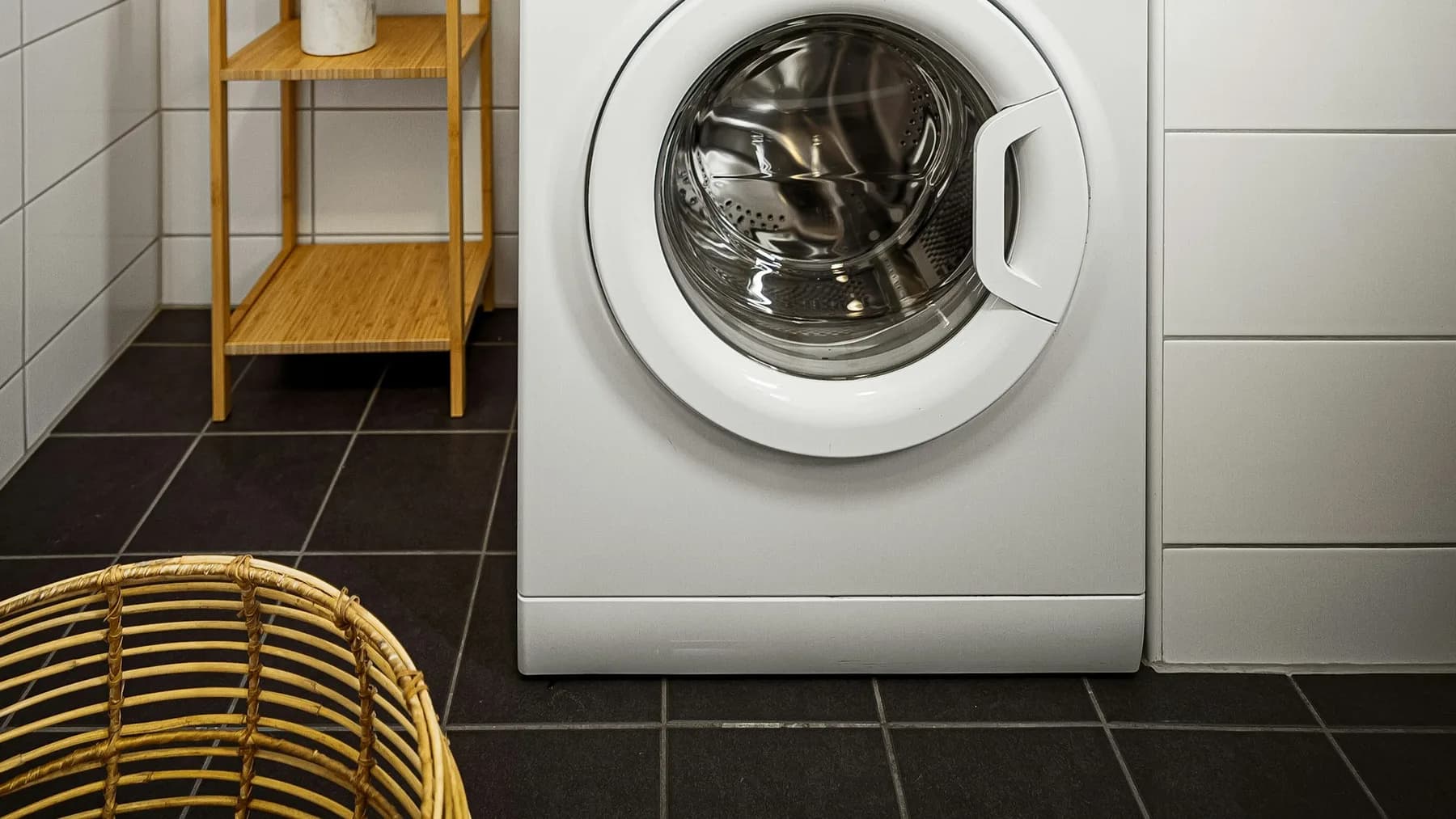 White front-loading washing machine standing on dark floor tiles beside a wicker laundry basket in a tiled utility room