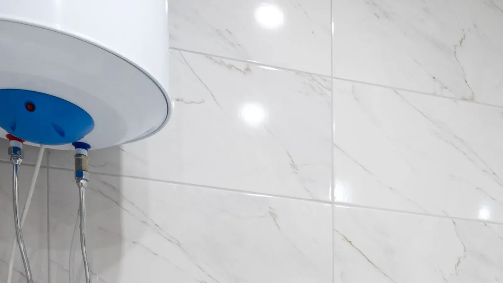 A wall-mounted water heater with two flexible hose connections against white marble-effect tiles in a Dubai bathroom
