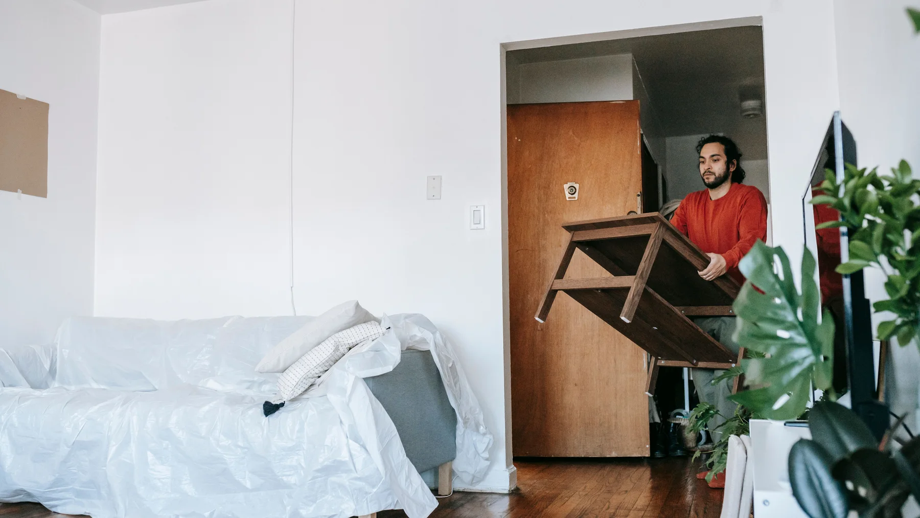 A man in a red jumper carrying a dark wooden table through an internal doorway of an apartment, past a sofa covered in protective plastic sheeting