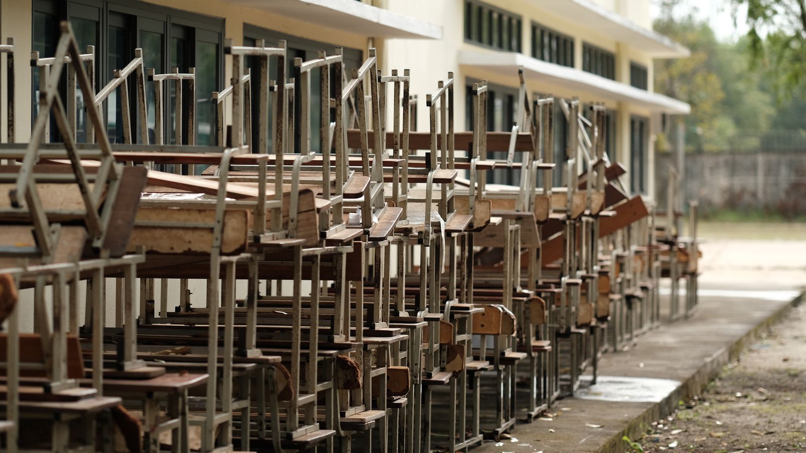 Rows of old wooden and metal desks and chairs stacked outside a building awaiting collection