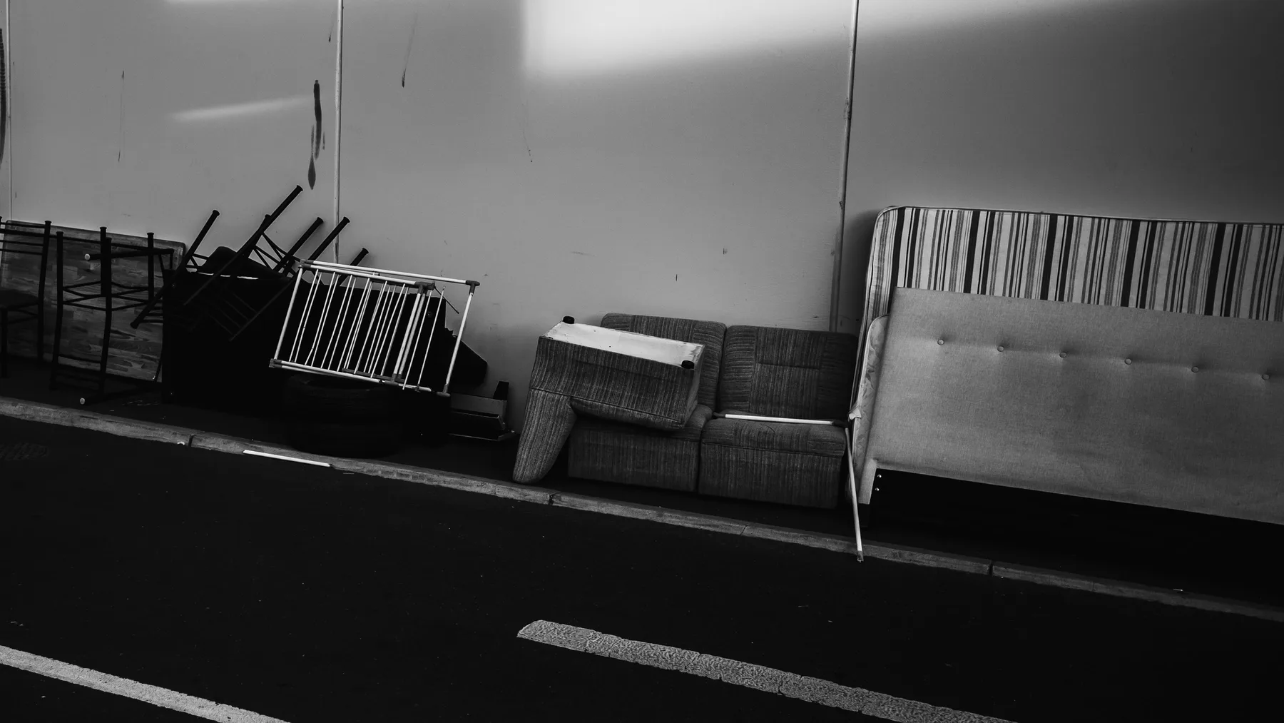 Discarded sofa, mattress base, bed frame and stacked chairs lined up along a wall at the edge of a car park