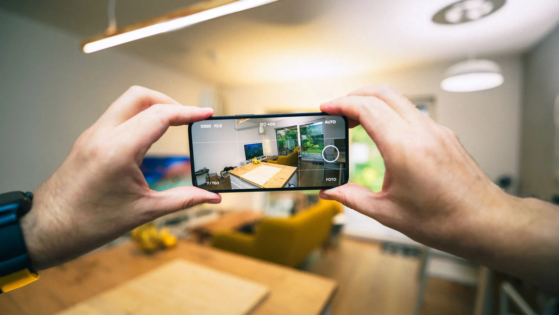 Two hands holding a smartphone in camera mode, framing a living room and dining table through the screen