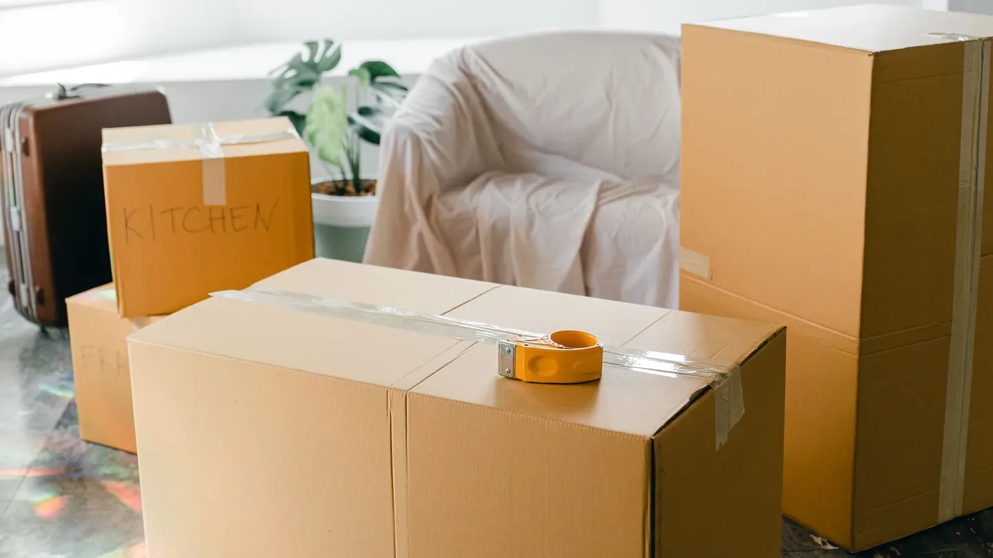 Sealed moving boxes and marked cartons beside a dust-sheeted armchair and suitcase in a room