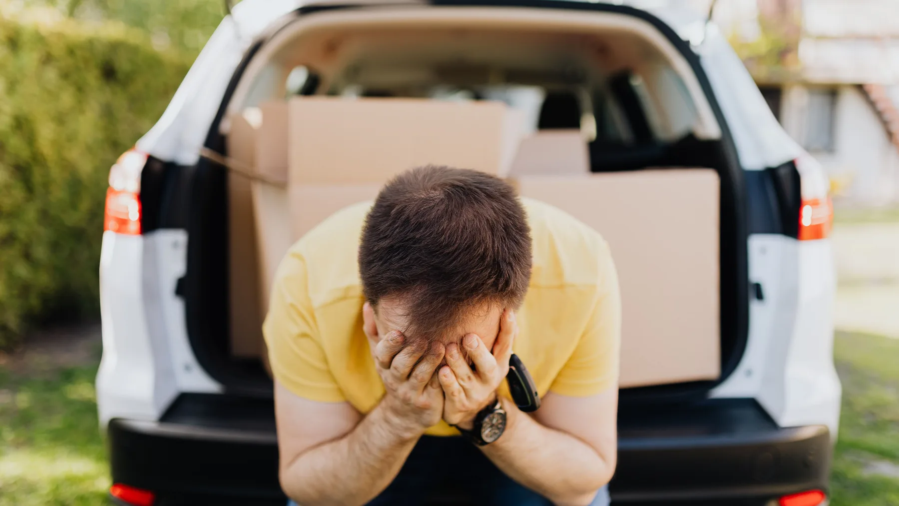 A frustrated man in a yellow shirt sits at the open boot of a white car loaded with cardboard boxes, holding his head in his hands