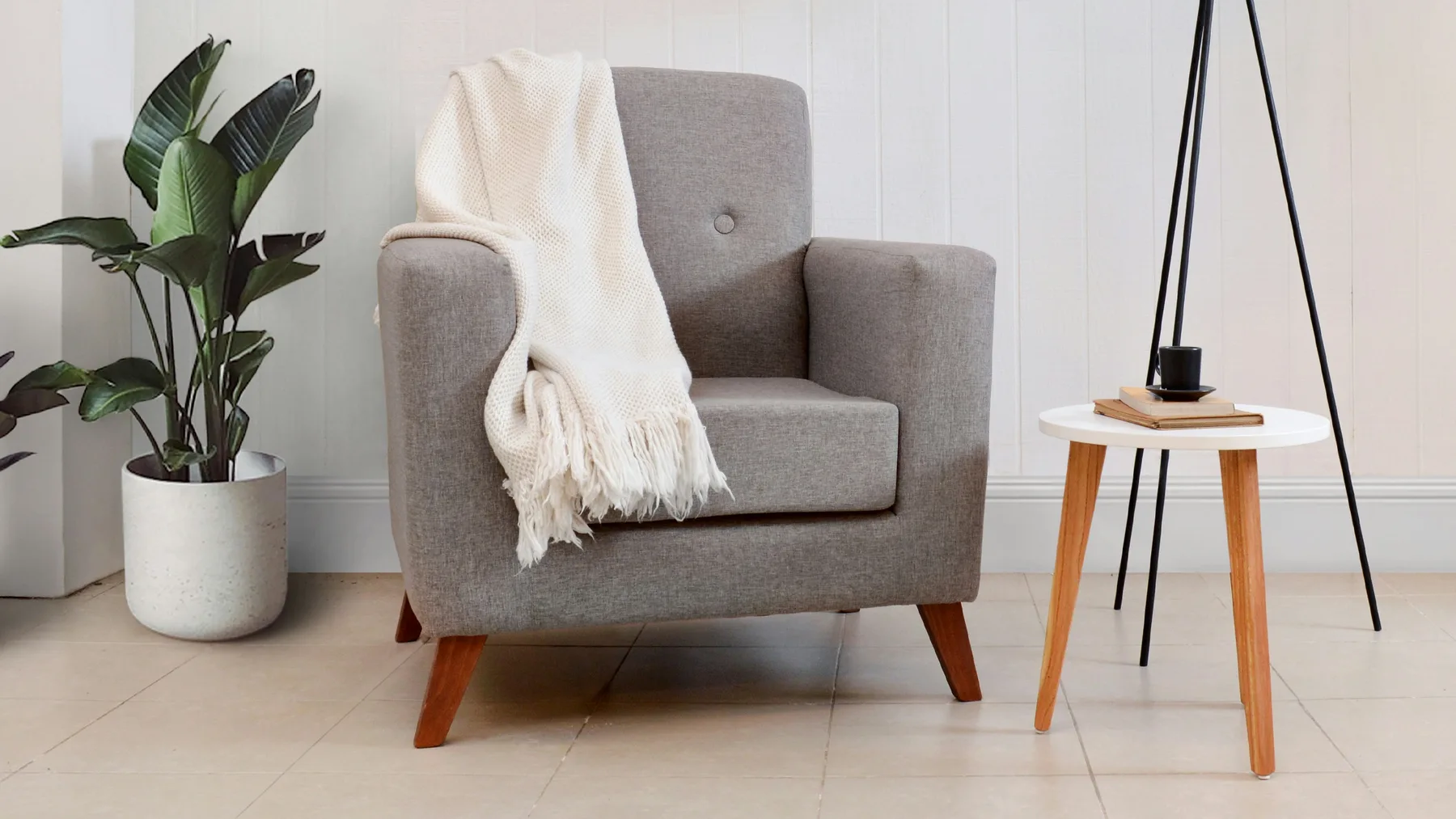 A grey upholstered armchair with a cream knitted throw, beside a potted plant and a small side table on tiled flooring