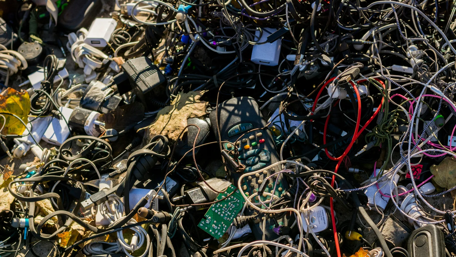 Large heap of tangled charger cables, power adaptors, earphones and two remote controls awaiting e-waste processing