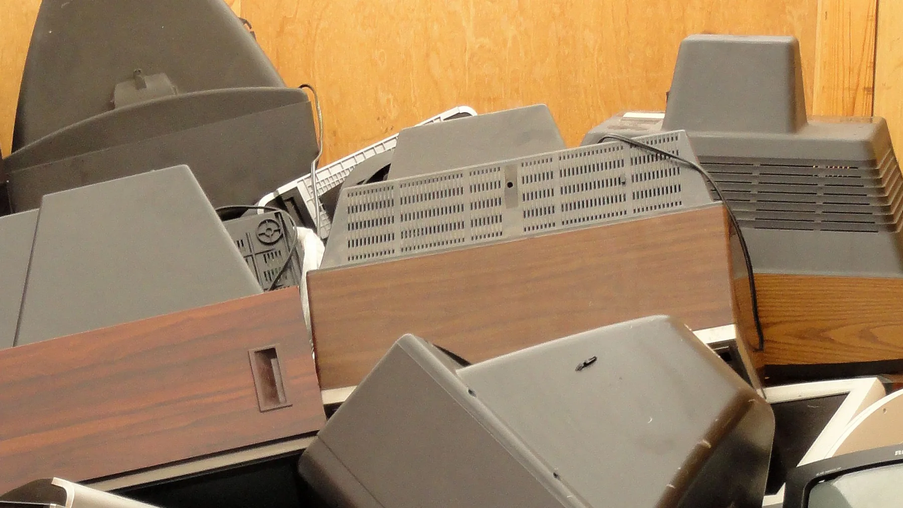 A large pile of old cathode-ray tube televisions and computer monitors stacked against a plywood wall, seen mostly from behind
