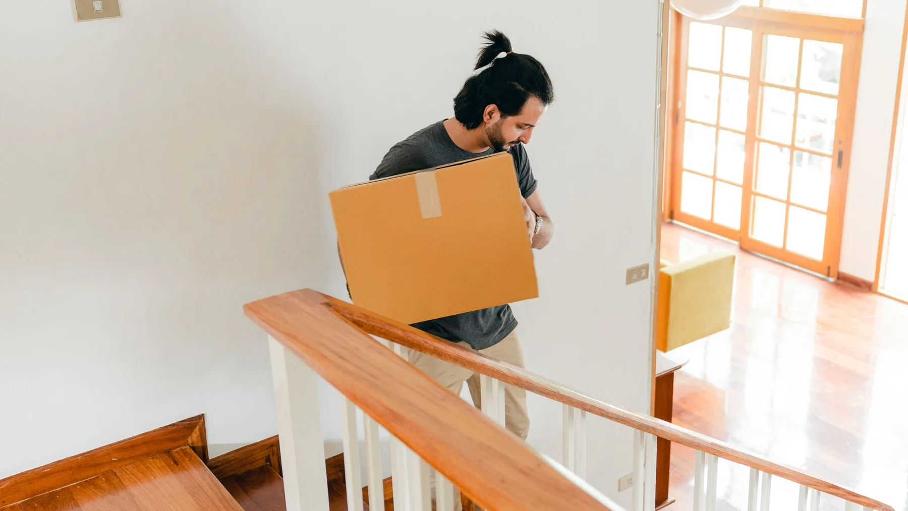 Man carrying a large sealed cardboard box down an indoor staircase past a wooden handrail