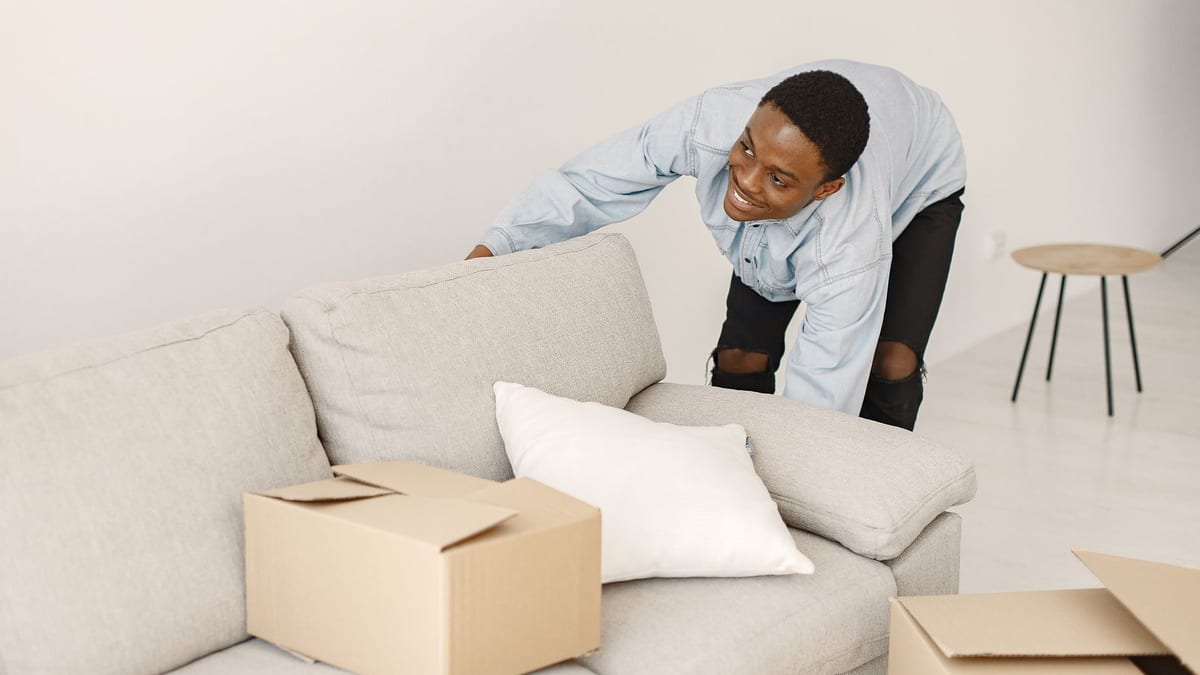 Man in a denim shirt lifting a grey sofa cushion in a bright room, with cardboard moving boxes on the floor in front of it