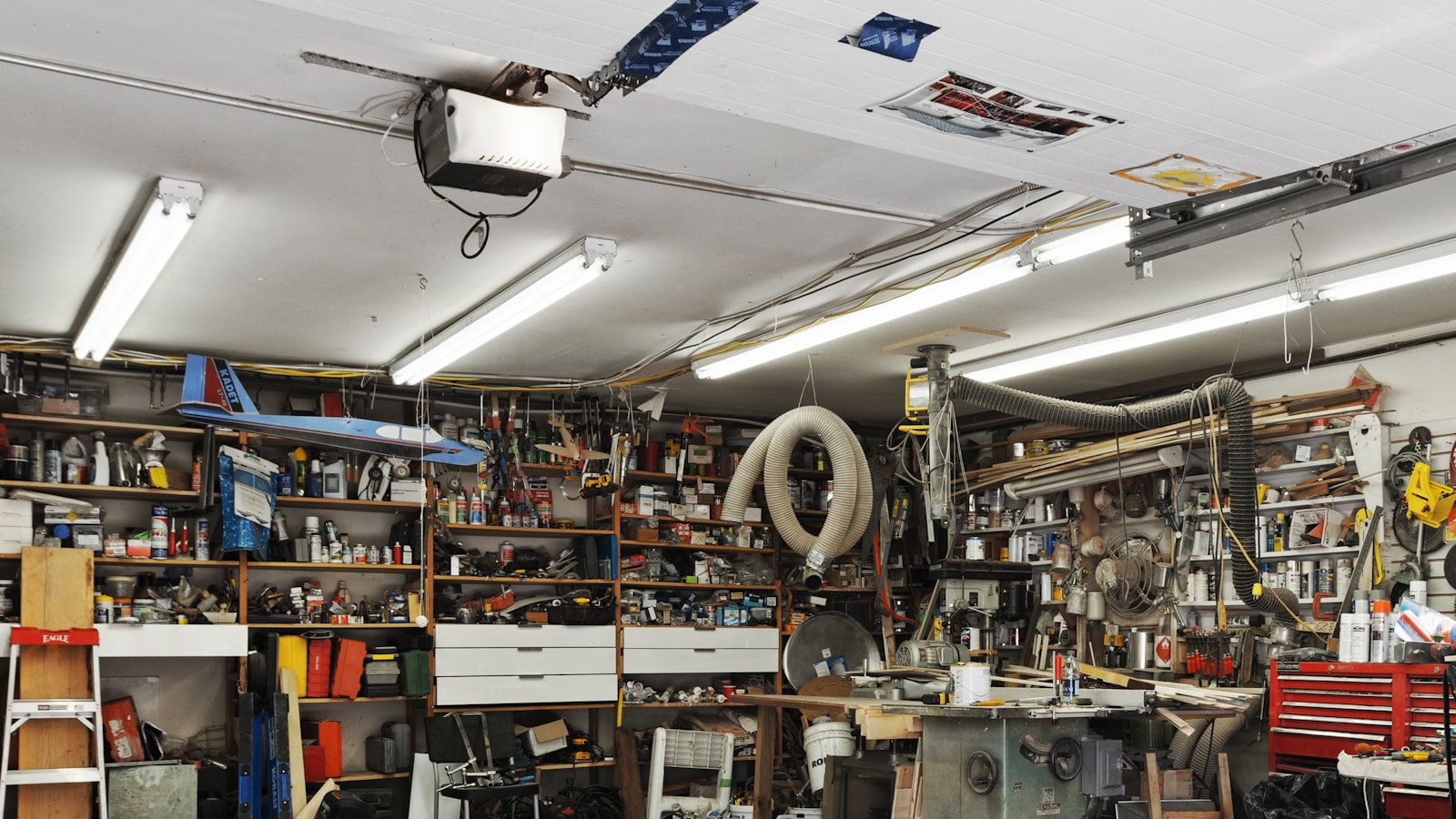 Cluttered garage with shelves of tools, boxes and stored items lining the walls above a concrete floor