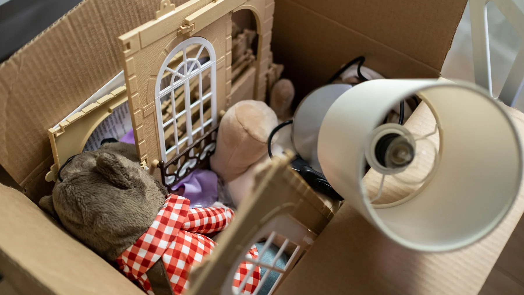 Open cardboard box holding a soft toy, a plastic toy house, a lampshade and tangled cable