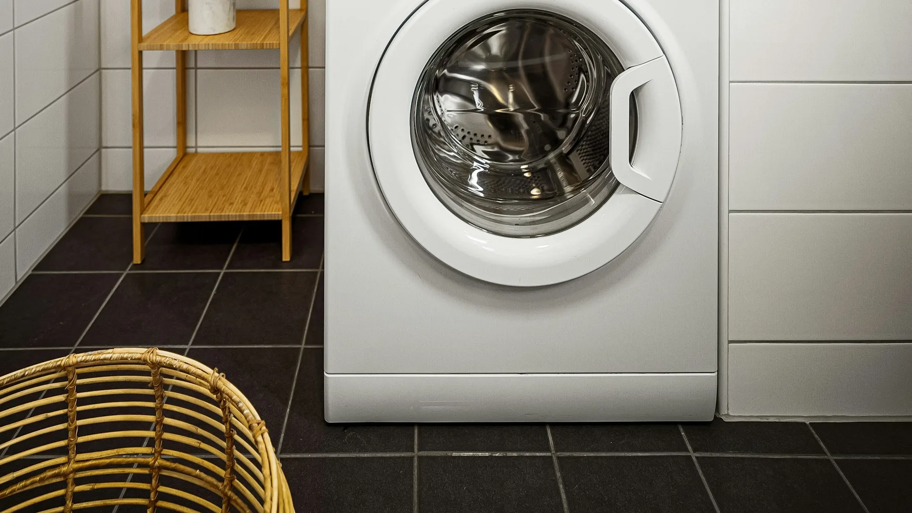 White front-loading washing machine standing on dark floor tiles beside a wicker laundry basket in a tiled utility room
