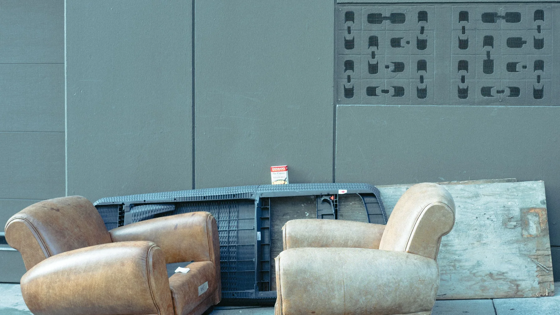 Two worn leather armchairs, a dark plastic frame and a stained timber board left against a plain grey wall on a paved pavement