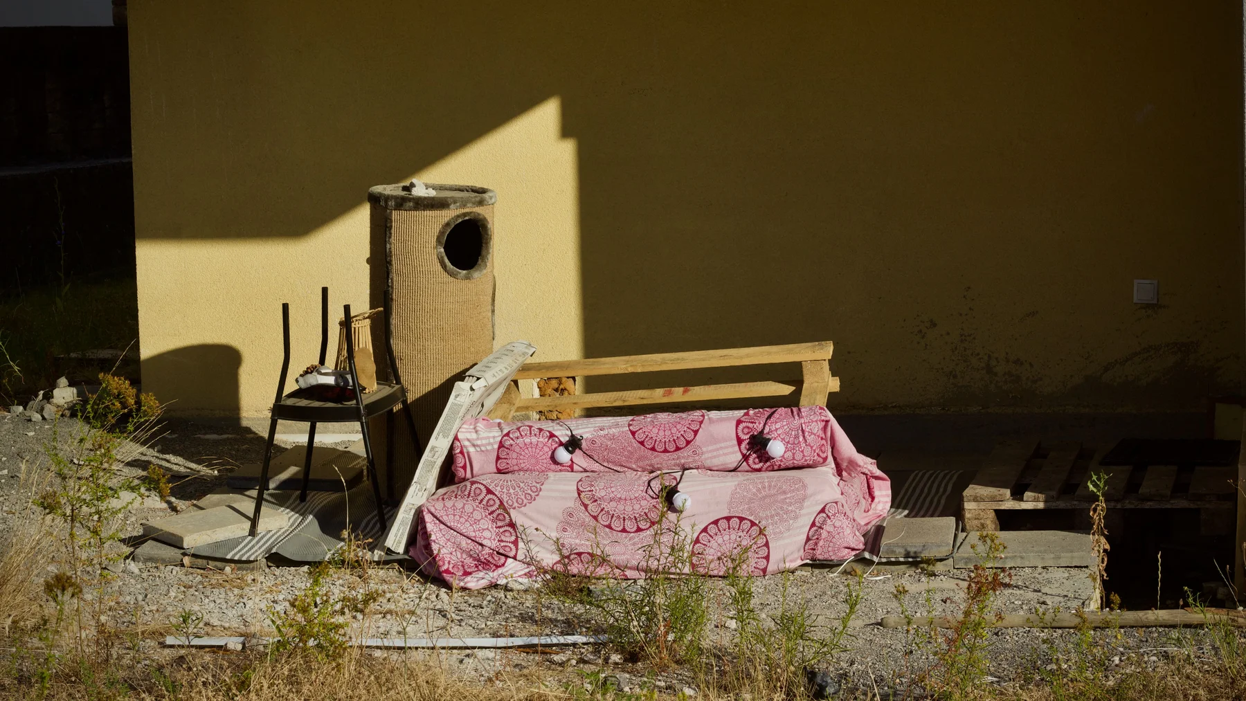 A fabric-covered bench seat, a metal chair and a fabric-wrapped column dumped against a sand-coloured wall on dry stony ground in strong sunlight