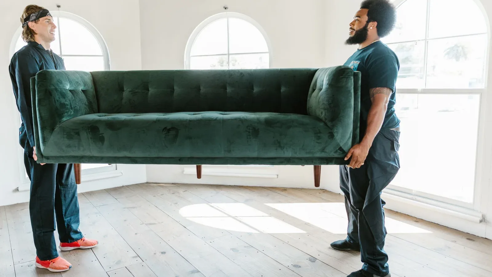 Two removal crew members carrying a green velvet sofa across a bright room with arched windows and a wooden floor