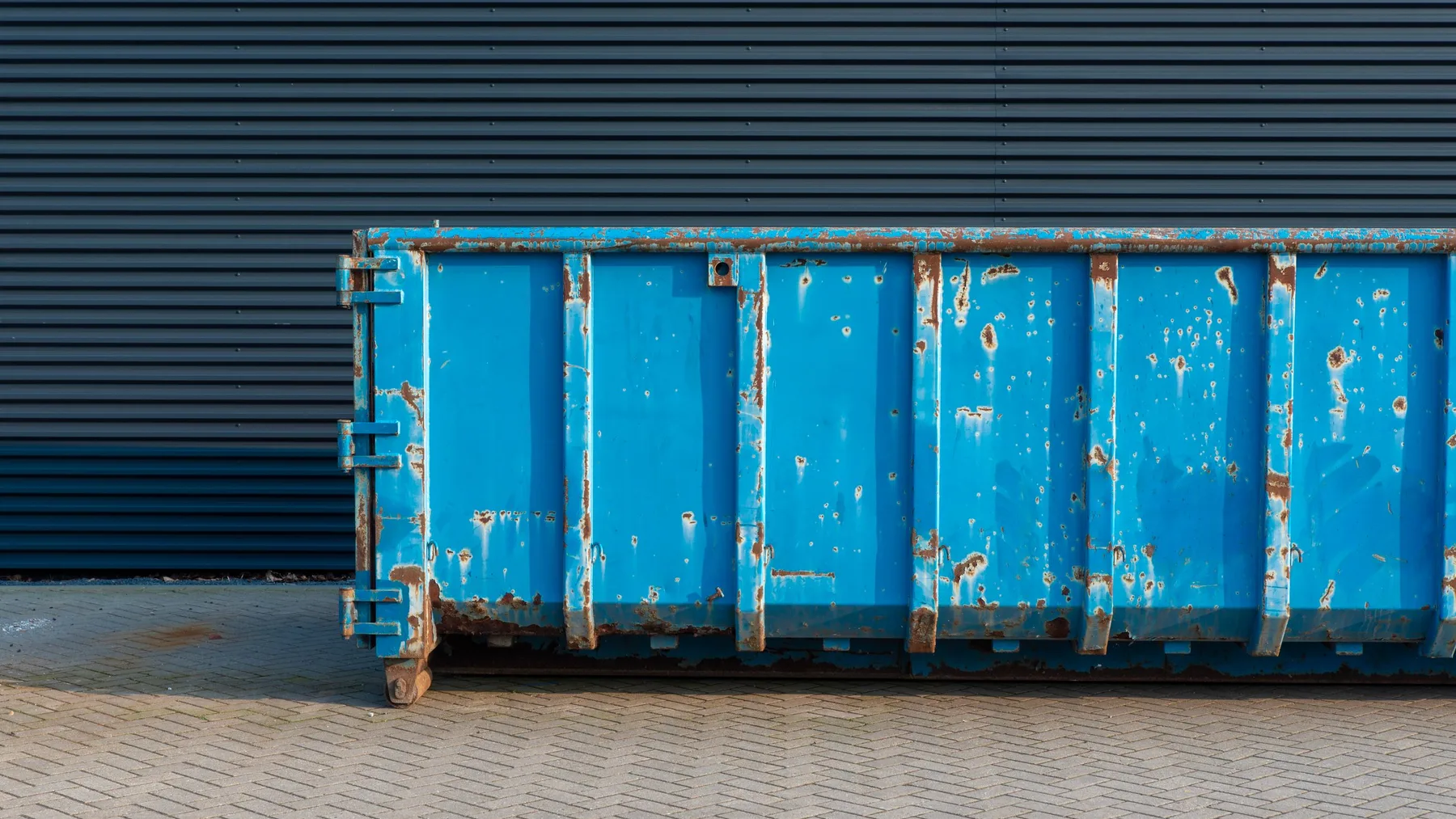 An empty blue steel waste container standing on block paving against a dark corrugated metal wall
