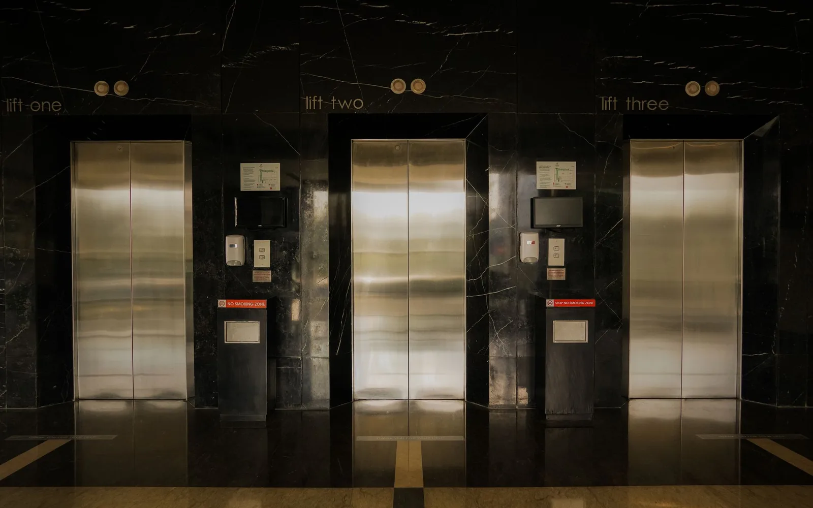 Three closed lift doors in a dark marble lobby of a residential building