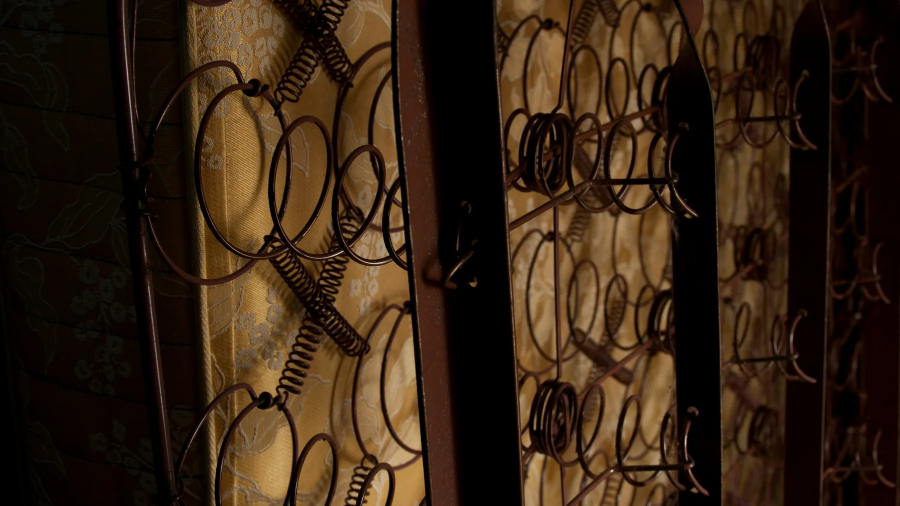 Rows of coiled metal springs and a wire frame exposed inside an old mattress with yellowed patterned fabric behind them