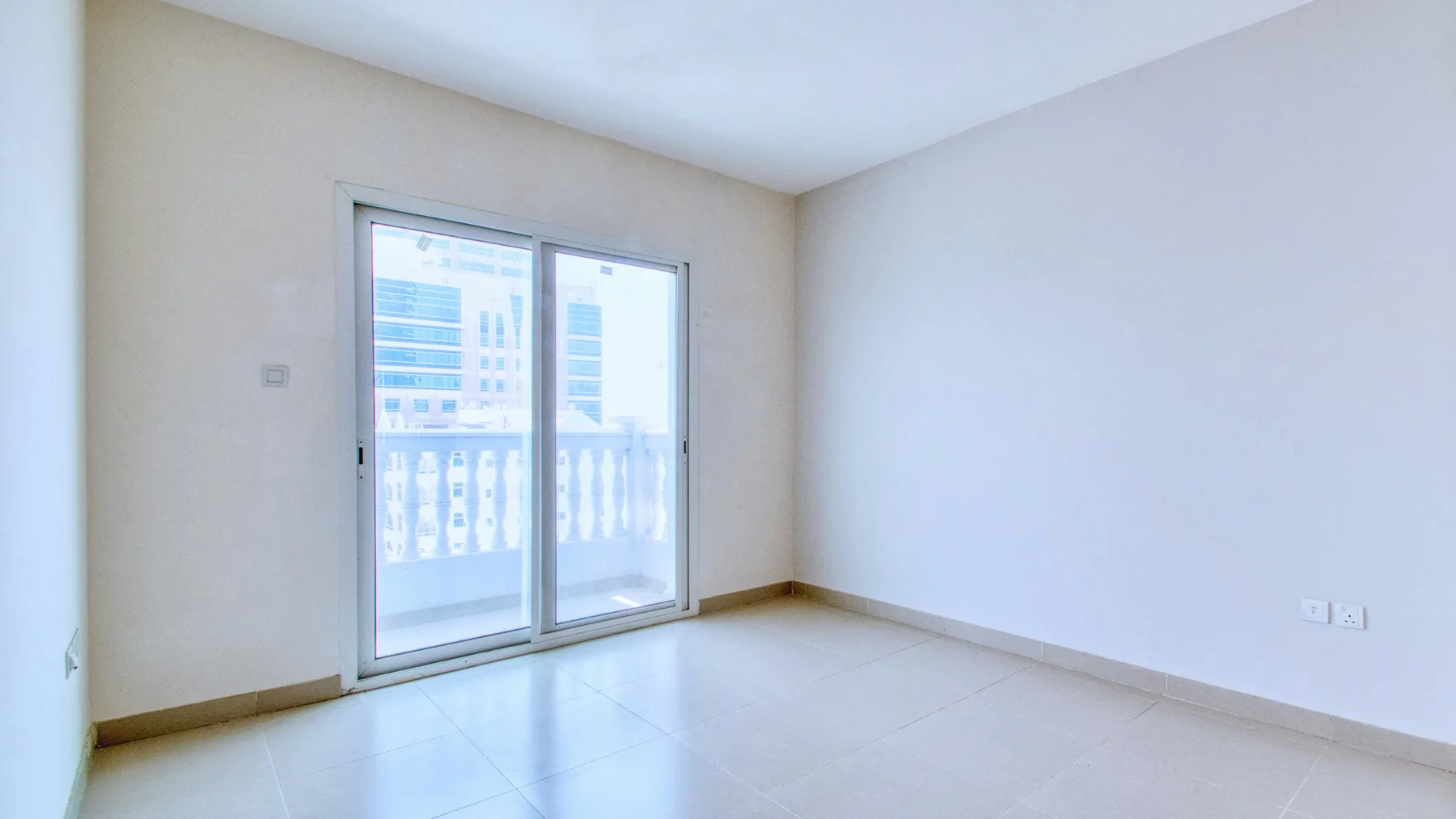 Bare apartment room with tiled floor and a sliding balcony door looking out onto neighbouring towers