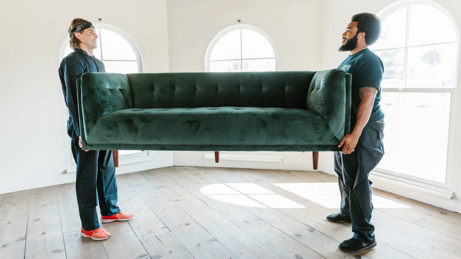 Two crew members in dark work overalls lifting a green velvet sofa together in a bright empty room with arched windows
