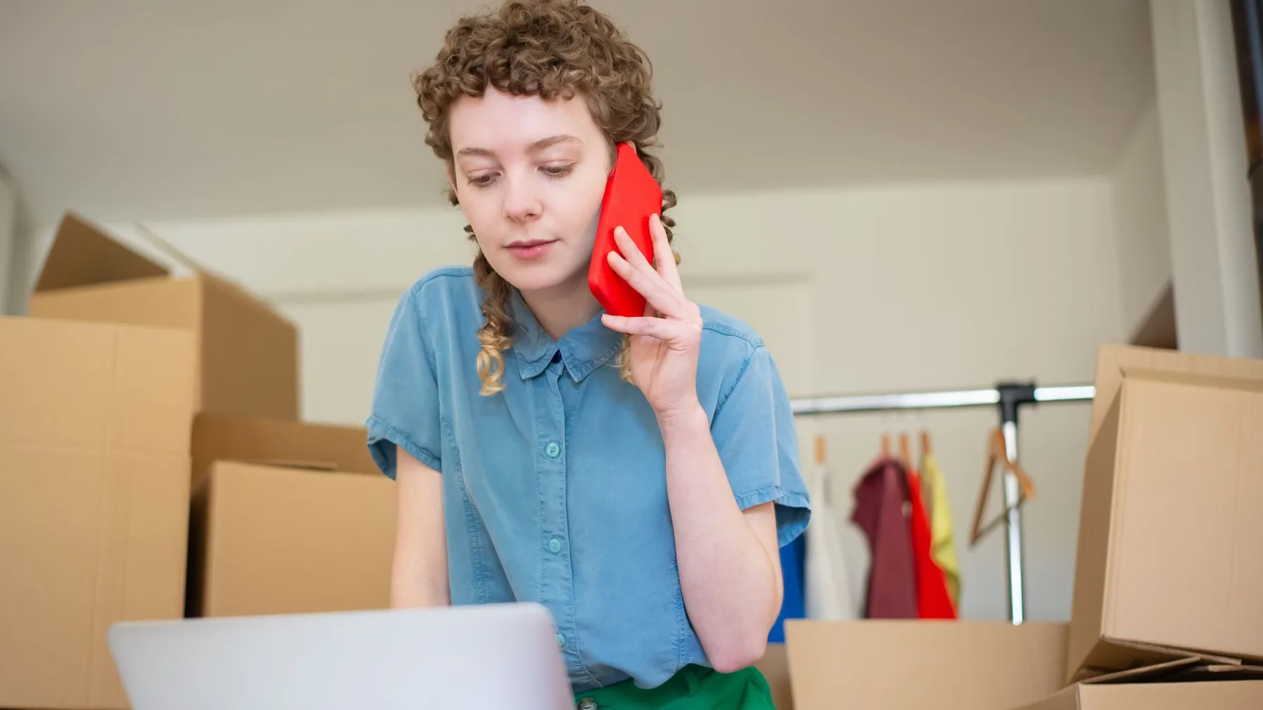 Woman holding a red phone to her ear while looking at a laptop, surrounded by stacked cardboard boxes
