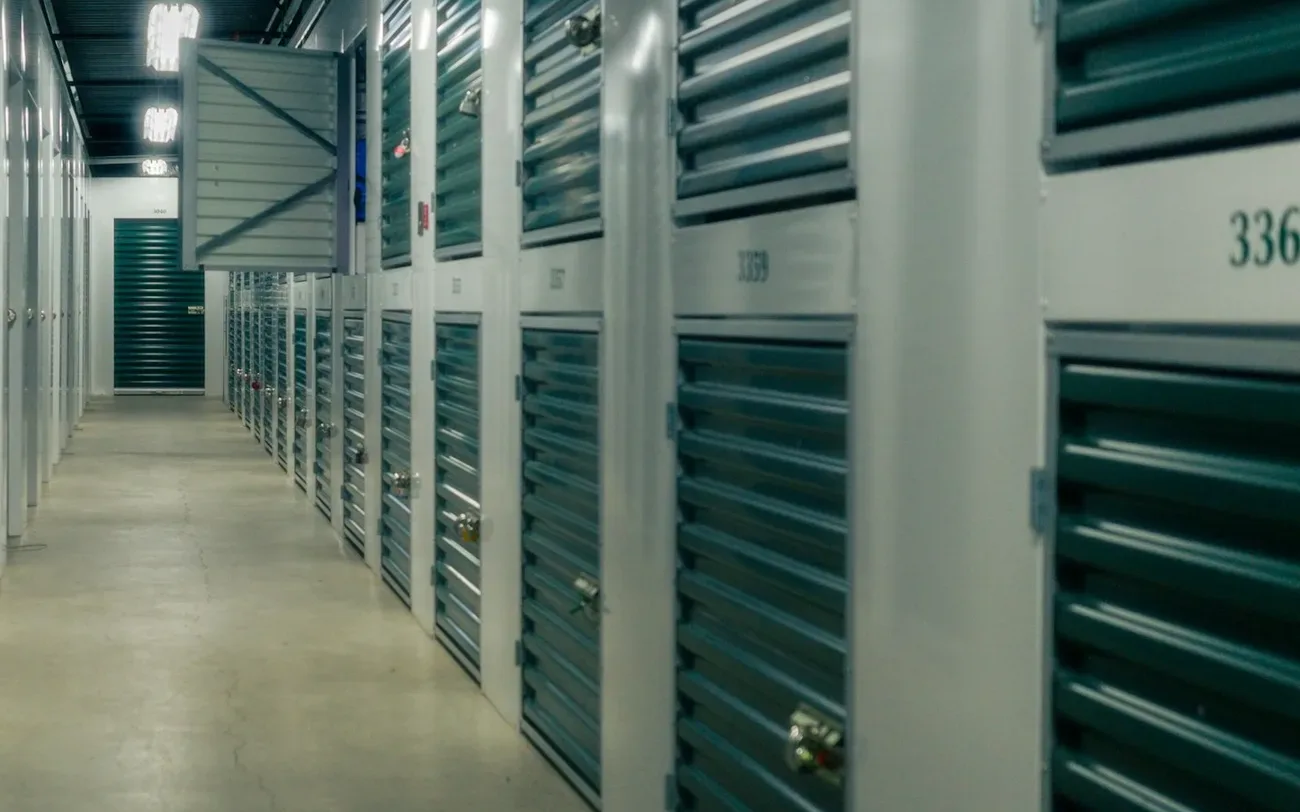 Long indoor self-storage corridor lined with numbered units with green roller shutters and padlocks