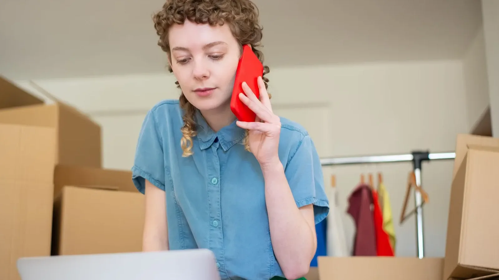 Woman on a red phone call checking a laptop among packed cardboard boxes and a clothes rail