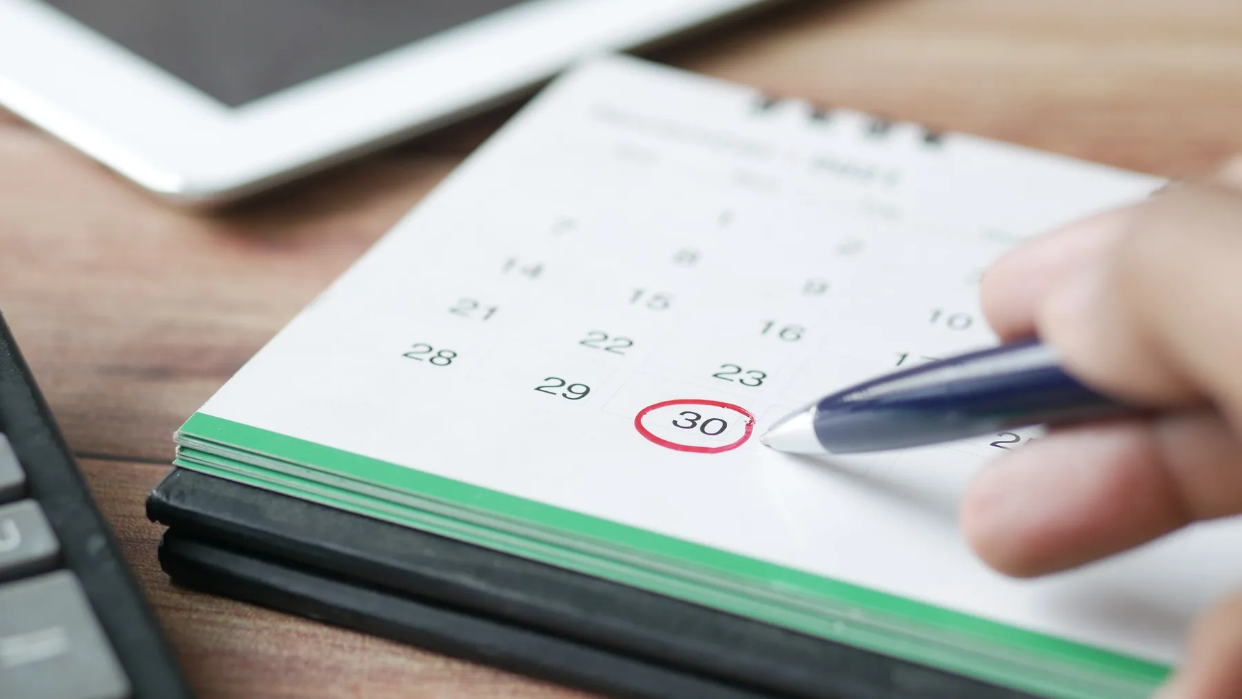 A hand circling a date on a desk calendar with a pen, next to a keyboard and tablet