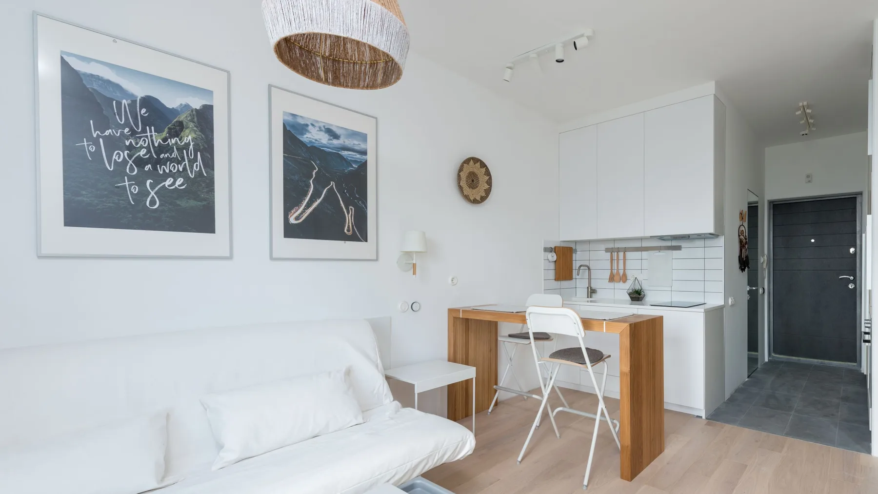 Compact studio apartment with a white sofa bed, narrow wooden bar table and a kitchenette with overhead cupboards beside the entrance door