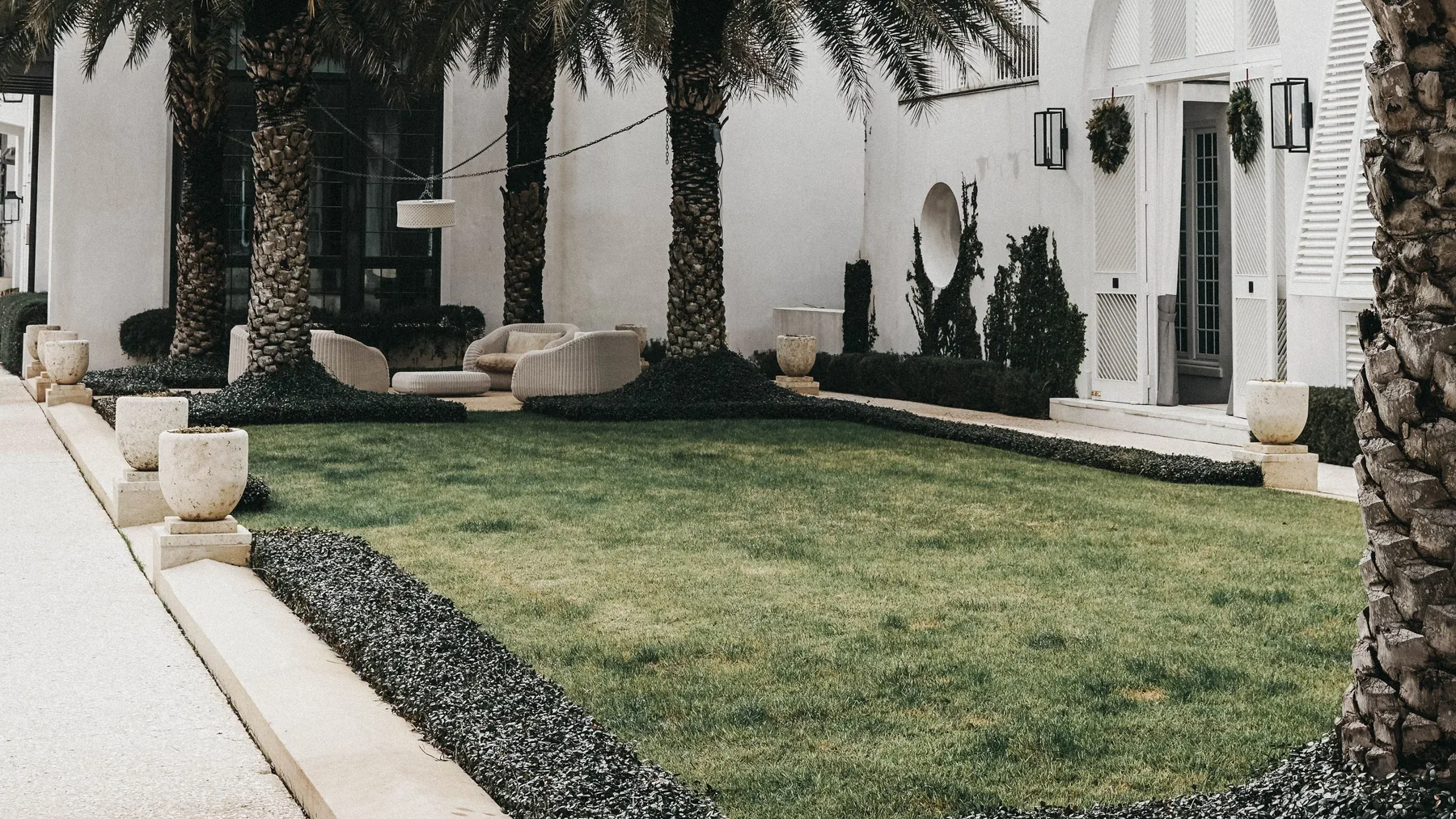 Villa courtyard with date palms, clipped hedging and rattan outdoor seating on a lawn beside a white rendered facade